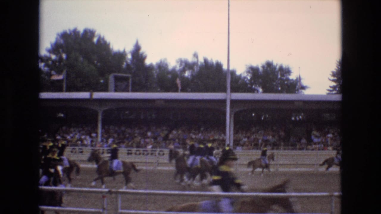 Mounted riders gallop past a crowded grandstand with track railings in the foreground.