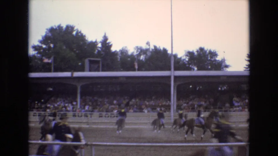 1973 Colorado Springs home-movie still showing horses and riders on a dirt track, with blurred foreground figures, a full grandstand, and trees beyond the roof.