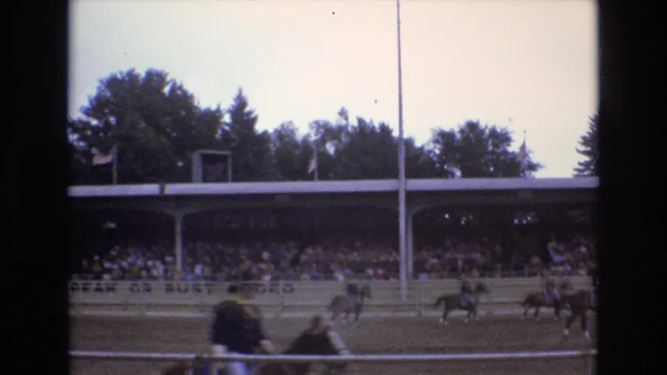 1973 Colorado Springs 8mm frame showing a rider on horseback moving along the track with a covered grandstand, railings, trees, and a tall pole behind the course.