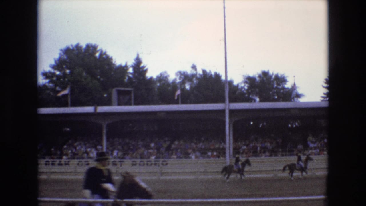 1973 Colorado Springs home-movie frame showing horseback riders in a dirt arena with a crowded covered grandstand, arena rail, trees, and flagpoles in the background.