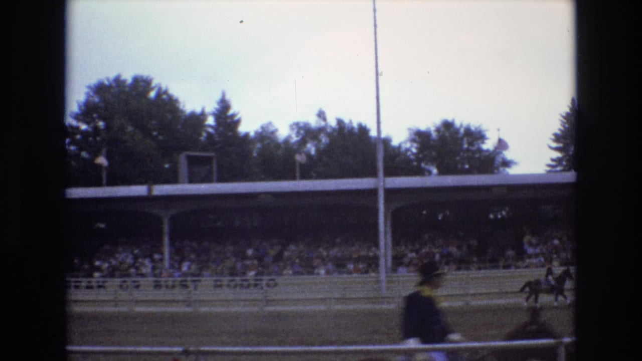 A blurred foreground rider passes while other horseback riders move near a crowded grandstand.