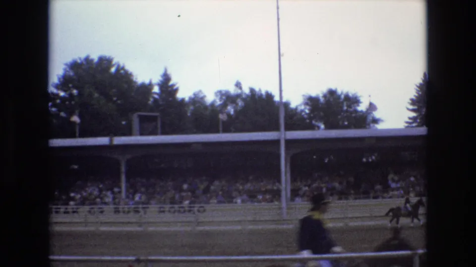 1973 Colorado Springs frame with a blurred rider crossing the foreground as other horses and riders move near the arena rail before a packed covered grandstand.