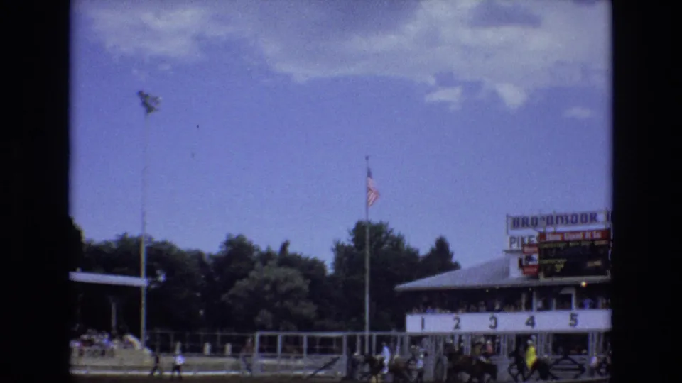 1973 Colorado Springs home-movie frame showing riders and horses near a starting gate area, with a sign reading Pike's Peak or Bust Rodeo and Mrs. Mora Ice Revue partly visible above the arena.