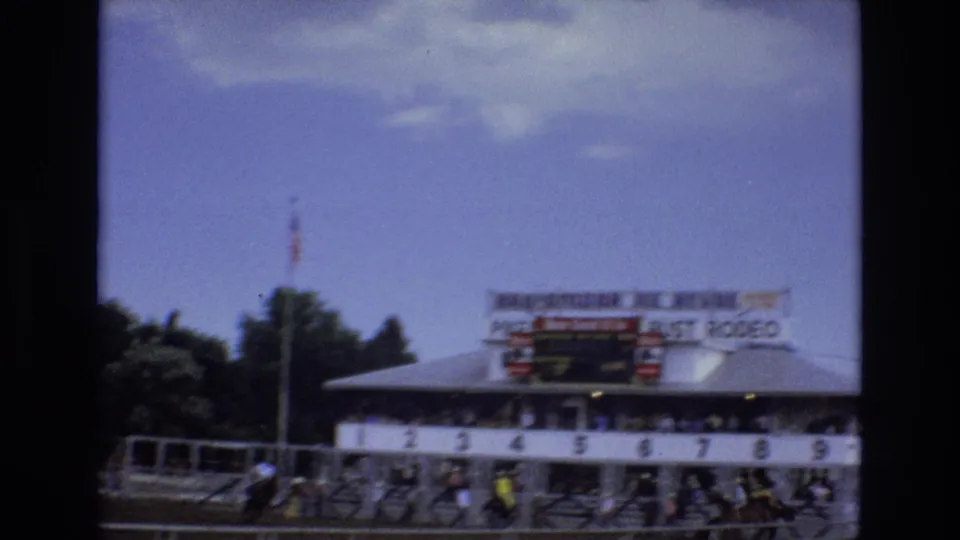 1973 Colorado Springs frame showing multiple horses and riders lined near numbered starting stalls under visible Pike's Peak or Bust Rodeo signage, with trees and sky behind the arena.