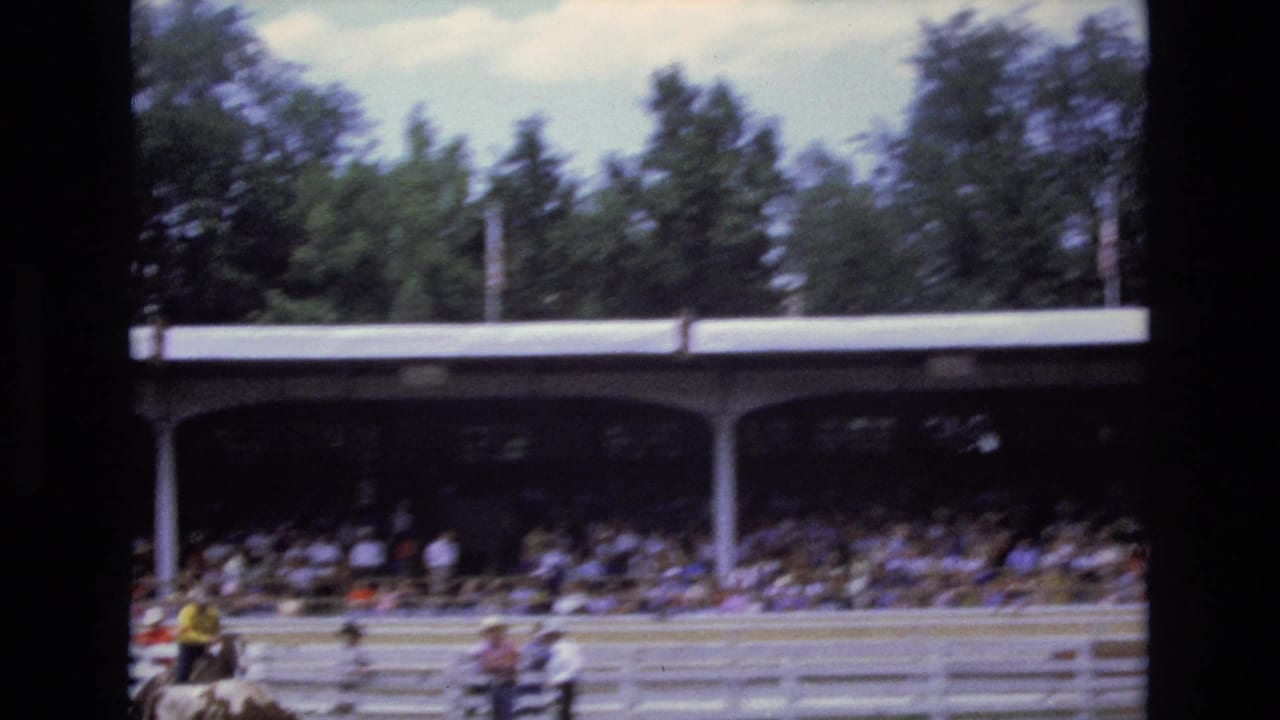 1973 Colorado Springs rodeo footage shows mounted riders moving across an arena in front of a crowded covered grandstand and trees.
