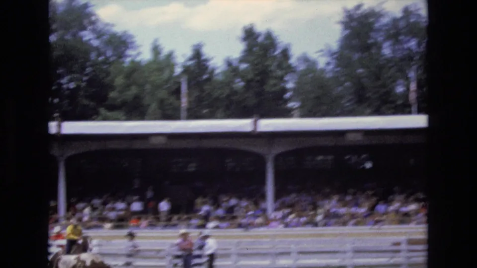 1973 Colorado Springs rodeo footage shows mounted riders moving across an arena in front of a crowded covered grandstand and trees.