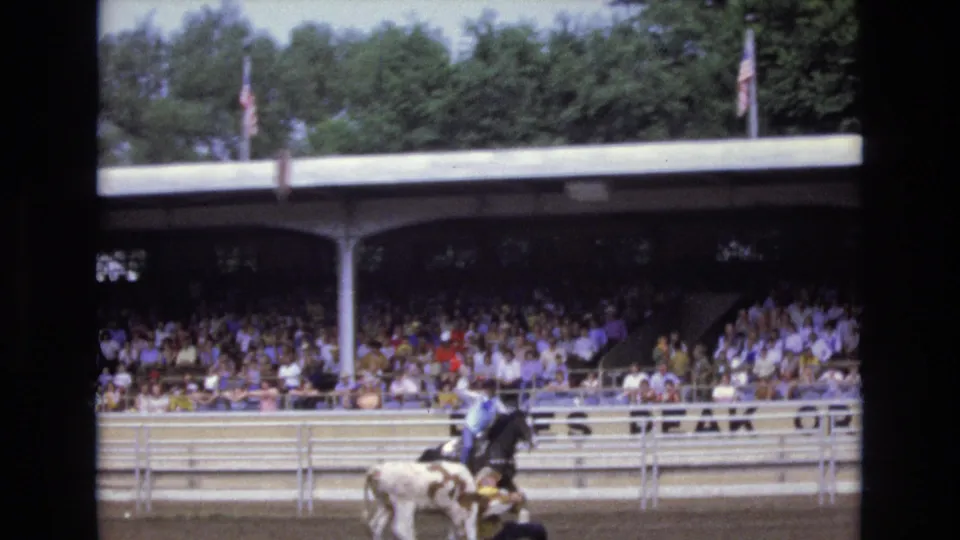 A rider and light-colored steer or calf pass the arena rail before a packed covered grandstand in the supplied 1973 Colorado Springs rodeo footage.