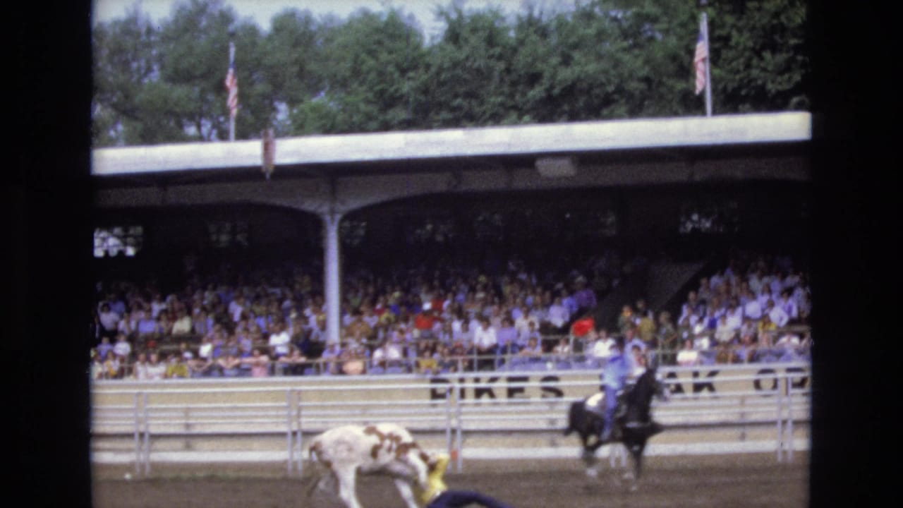 1973 Colorado Springs rodeo home-movie frame showing a white-and-brown bull in the arena, a rider on horseback nearby, and a large crowd watching from the covered stands.