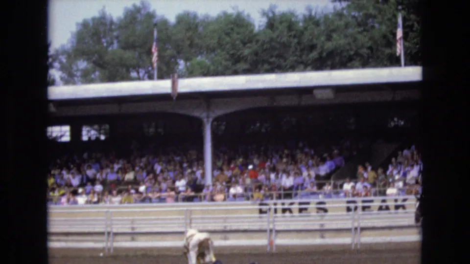 1973 Colorado Springs rodeo home-movie frame showing the bull near the foreground rail, a dense grandstand crowd, roof columns, flags, and partial Pikes Peak lettering.