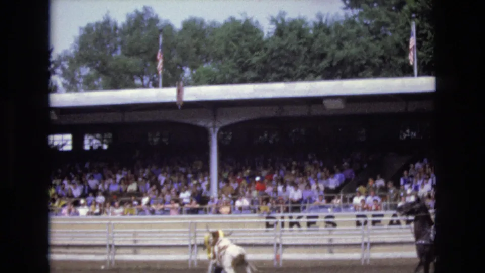 1973 Colorado Springs rodeo frame with the bull partly at the lower right edge, the grandstand crowd in the background, American flags above the roofline, and visible Pikes Peak lettering on the stands.