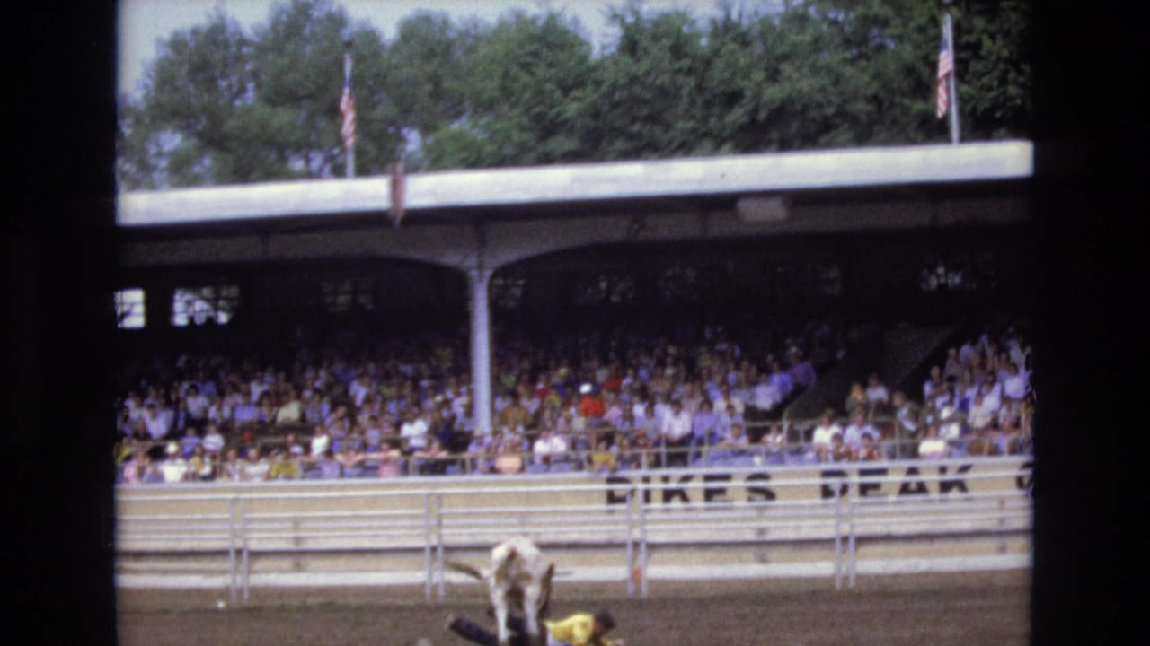 1973 Colorado Springs rodeo scene with a bucking bull partly visible at the bottom of the frame, spectators under covered stands, American flags, rail fencing, and partial venue lettering.