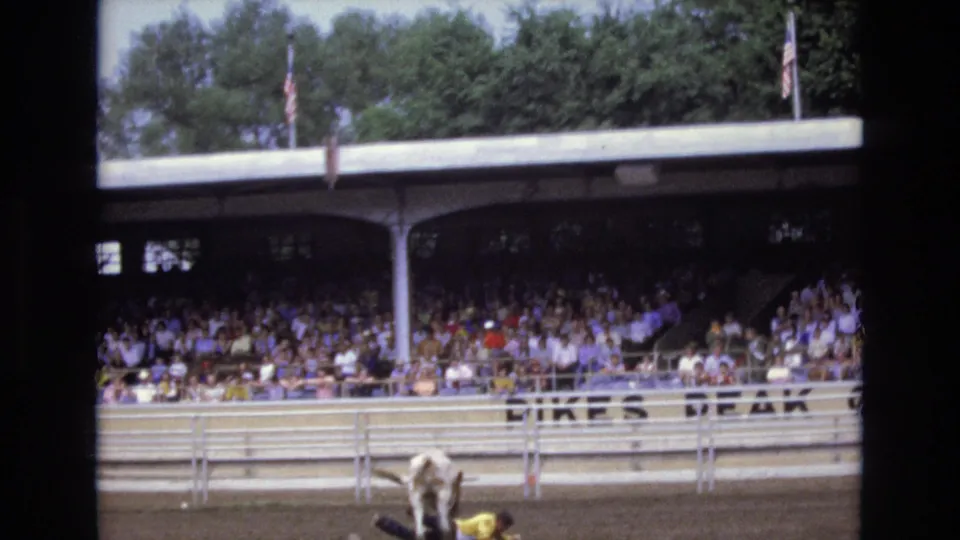 1973 Colorado Springs rodeo scene with a bucking bull partly visible at the bottom of the frame, spectators under covered stands, American flags, rail fencing, and partial venue lettering.
