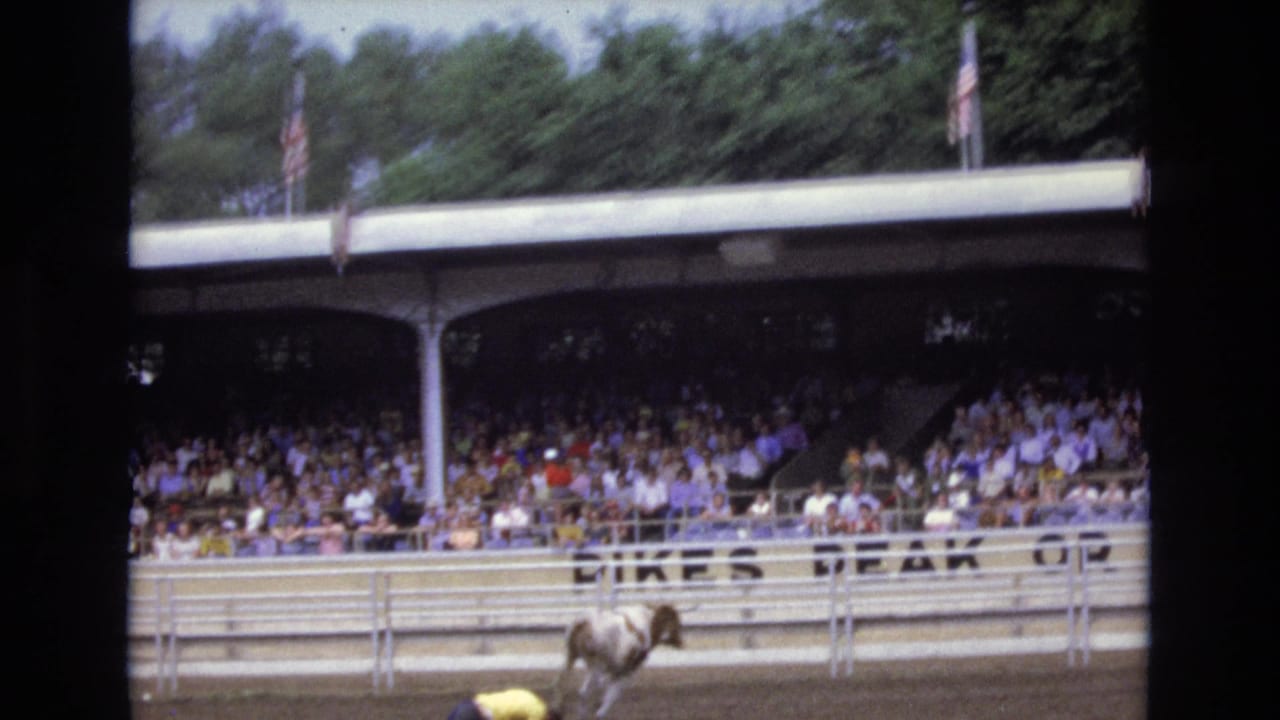 A bull or rider is partly visible near the bottom edge as the home-movie camera frames the covered rodeo stands, crowd, flags, arena rail, and partial lettering in Colorado Springs.