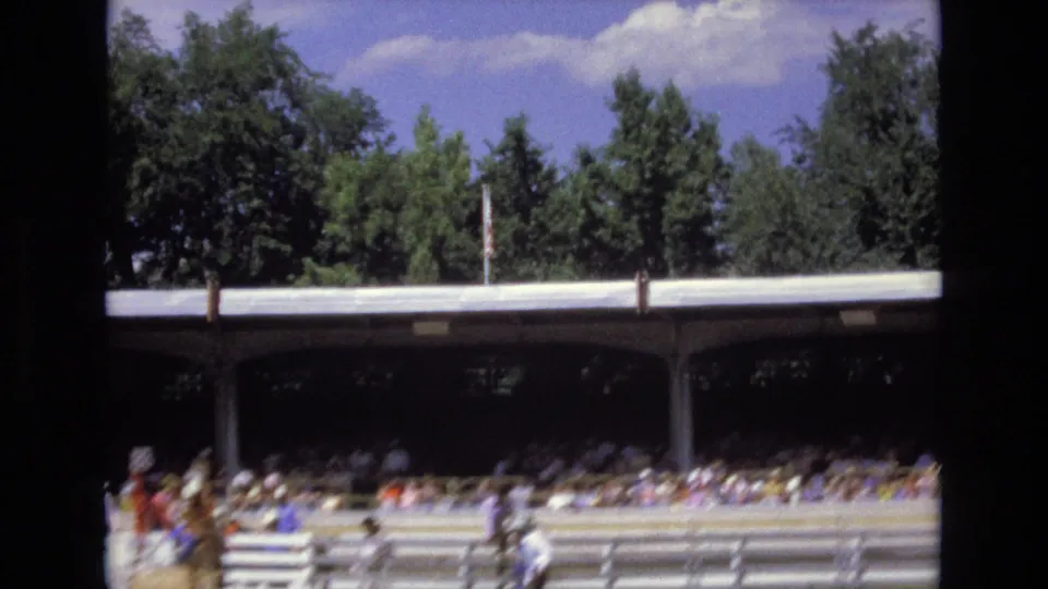 Home-movie view of the rodeo grounds with horseback riders or arena staff near the lower edge, a shaded spectator stand, rail fencing, trees, and blue sky.