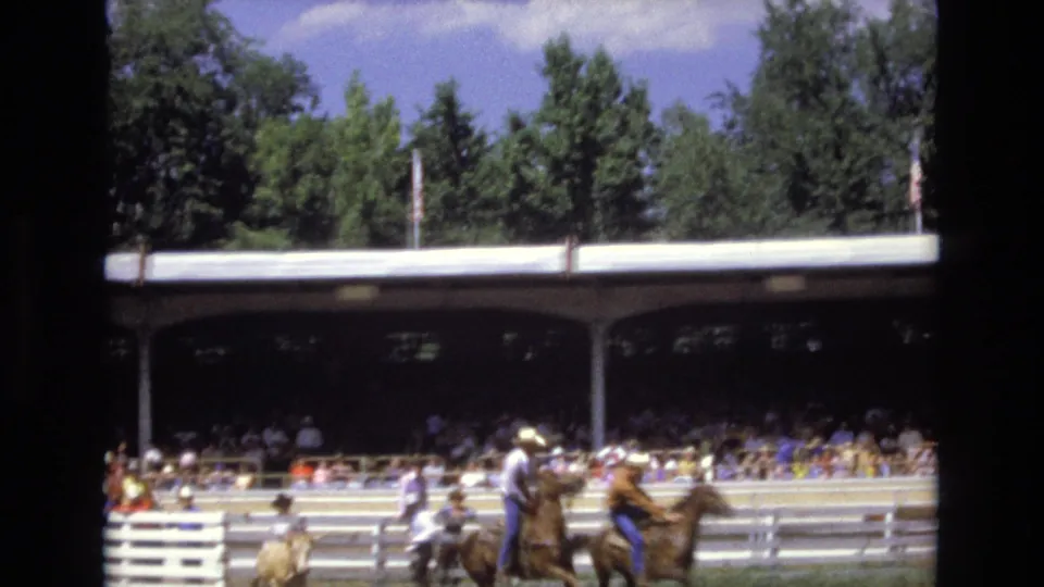 Riders on horseback and arena personnel cluster in the dirt ring at the Colorado Springs rodeo, with crowded covered bleachers, rail fencing, trees, and an American flag behind them.