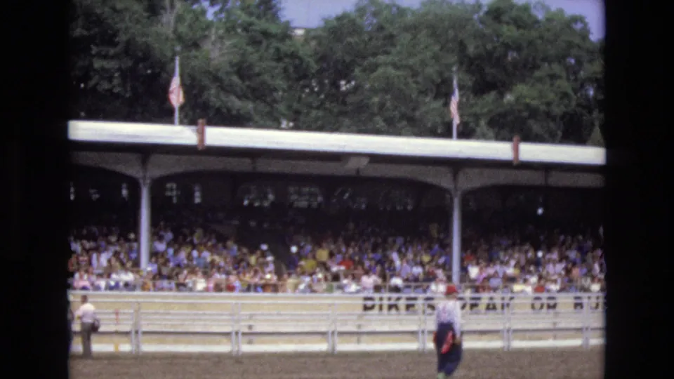 1973 Colorado Springs 8mm frame of a covered grandstand crowded with spectators, flags above the roof, and a dirt arena bordered by white rails in the foreground.