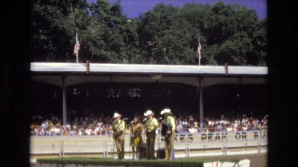 1973 Colorado Springs home-movie frame showing a small group of musicians or performers in cowboy hats near the arena rail with covered bleachers and spectators behind them.