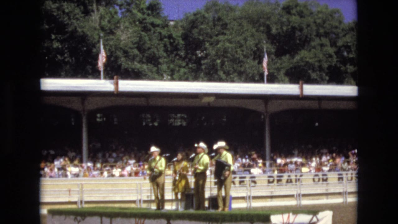 In 1973 Colorado Springs, a country band in coordinated western-style clothing performs outdoors for a large seated audience under a shaded pavilion.