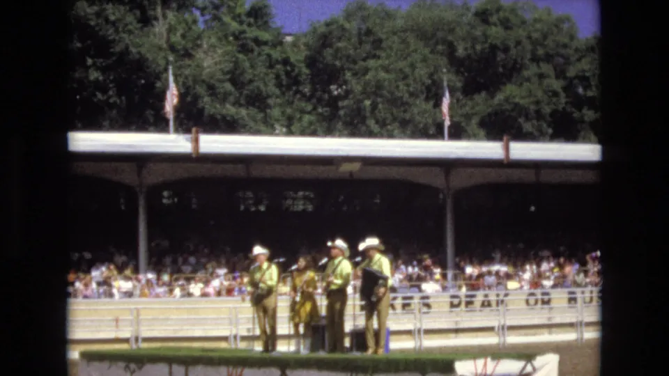 In 1973 Colorado Springs, a country band in coordinated western-style clothing performs outdoors for a large seated audience under a shaded pavilion.
