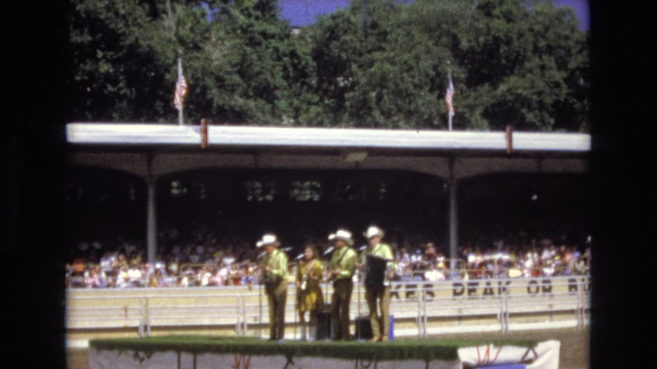 Musicians in cowboy hats stand with instruments near a microphone as the audience watches.