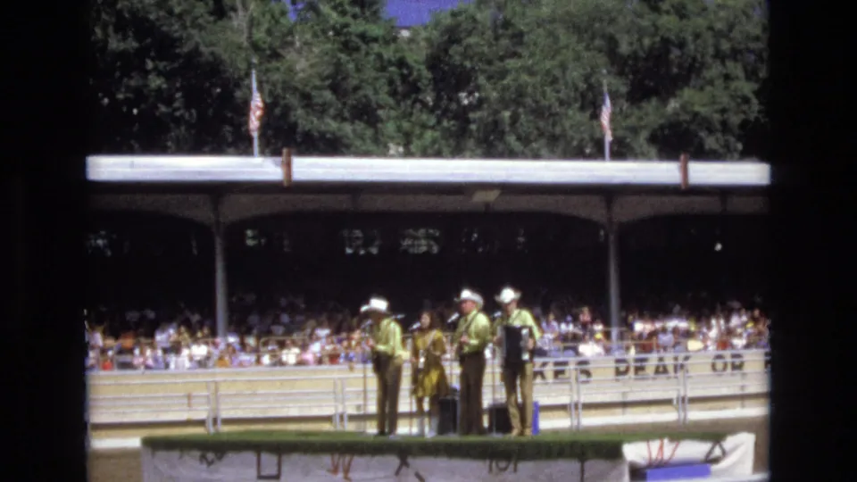 The closing frame shows the 1973 Colorado Springs outdoor country band from a distance, framed by railings, flags, trees, and a large shaded audience.