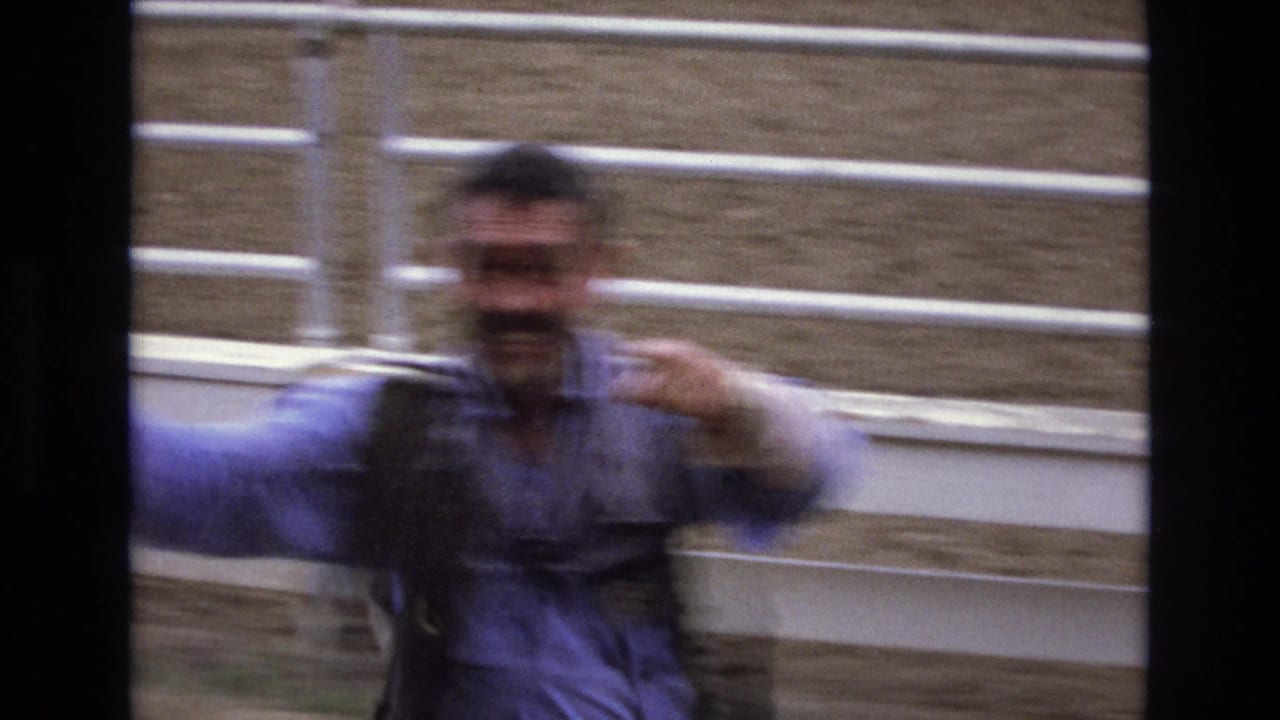 In 1973 Colorado Springs rodeo footage, a blurred man in a blue shirt and vest gestures close to white arena fencing as the handheld camera tracks his movement.