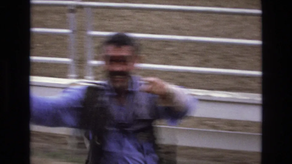 In 1973 Colorado Springs rodeo footage, a blurred man in a blue shirt and vest gestures close to white arena fencing as the handheld camera tracks his movement.