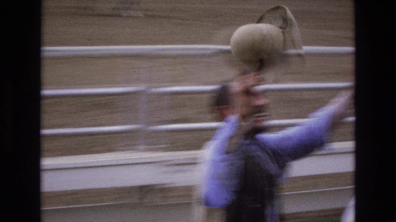 Man raises a cowboy hat beside white fencing in a rodeo arena.