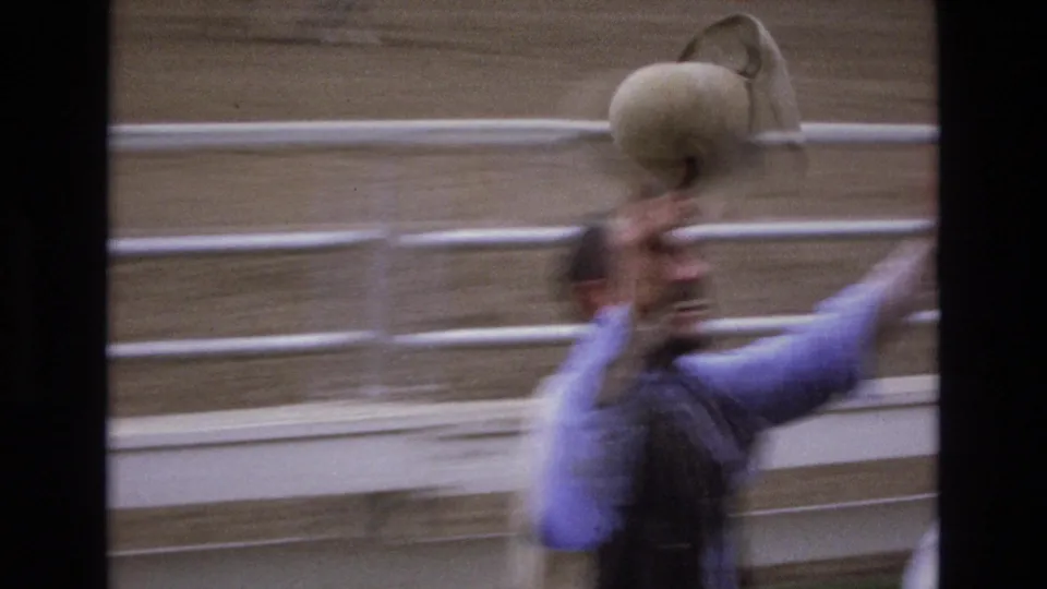 A man at the Colorado Springs rodeo raises a cowboy hat beside white arena rails, with his face partly blurred by fast handheld motion.