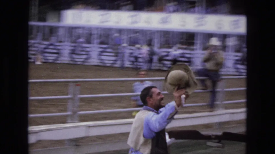 The celebrating man smiles and lifts a cowboy hat near the white rodeo fence, with grandstand seating and other arena figures visible in the background.