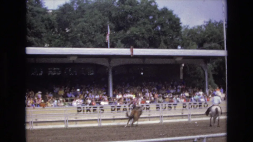A wider Colorado Springs rodeo view shows a dirt arena, white fencing, a covered grandstand full of spectators, partial arena signage, and riders on horseback.