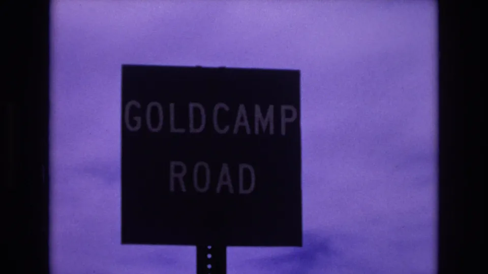 Colorado Springs 8mm frame with the Gold Camp Road sign framed lower in the shot, showing dark lettering, sign post, purple sky, and film shadowing.