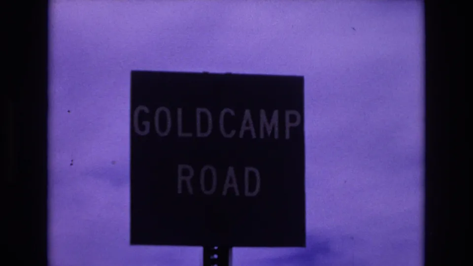1973 Colorado Springs home-movie frame showing a closer Gold Camp Road sign, with dark sign face, pale lettering, purple color shift, and visible 8mm grain.