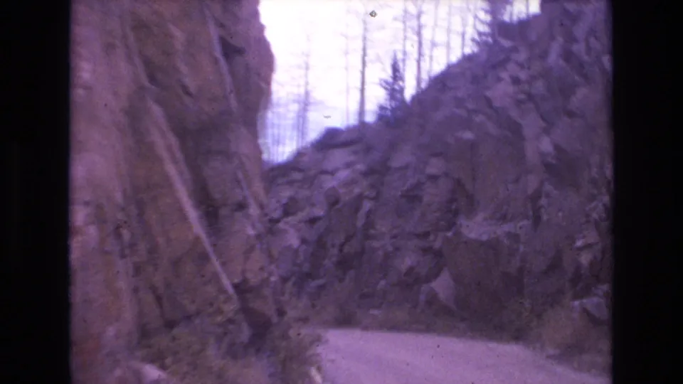 A narrow mountain road appears to curve through steep rocky walls near Colorado Springs, with sparse trees visible along the ridge in purple-shifted 8mm color.