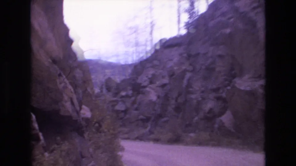 The home-movie view looks through a rocky roadside passage toward distant hills, with the road edge and cliff faces softened by 8mm grain and color fading.