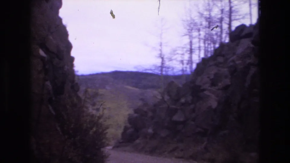 A rocky roadside opening near Colorado Springs looks out toward low distant hills, with sparse trees silhouetted against a pale sky in faded 8mm color.