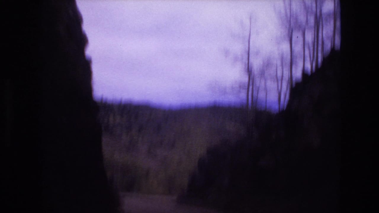 1973 Colorado Springs 8mm frame looking through dark rock formations toward a muted hillside, sparse trees, and an overcast purple-tinted sky.