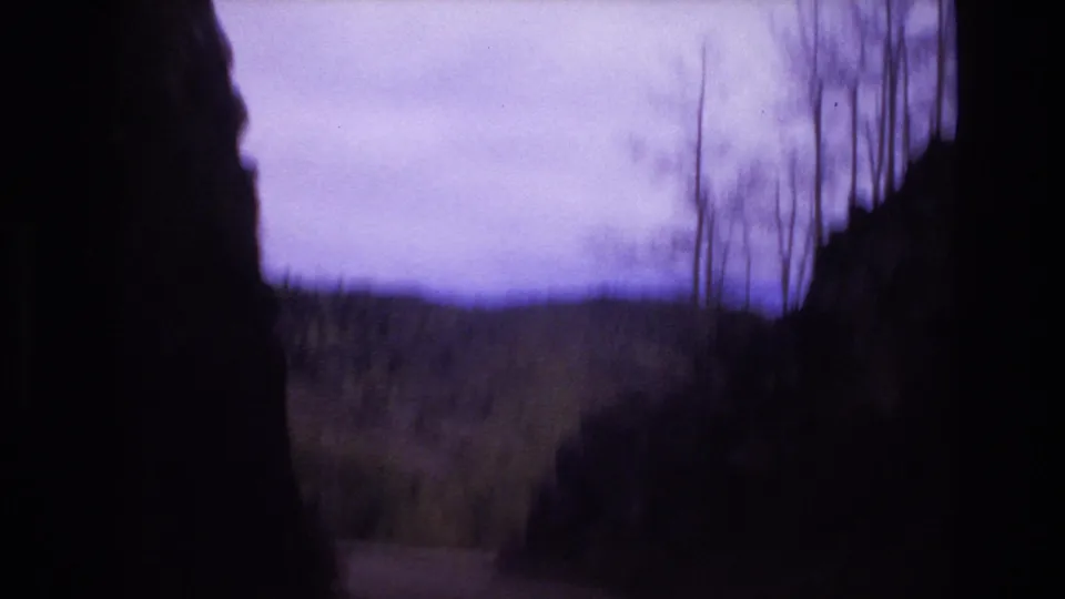 1973 Colorado Springs 8mm frame looking through dark rock formations toward a muted hillside, sparse trees, and an overcast purple-tinted sky.