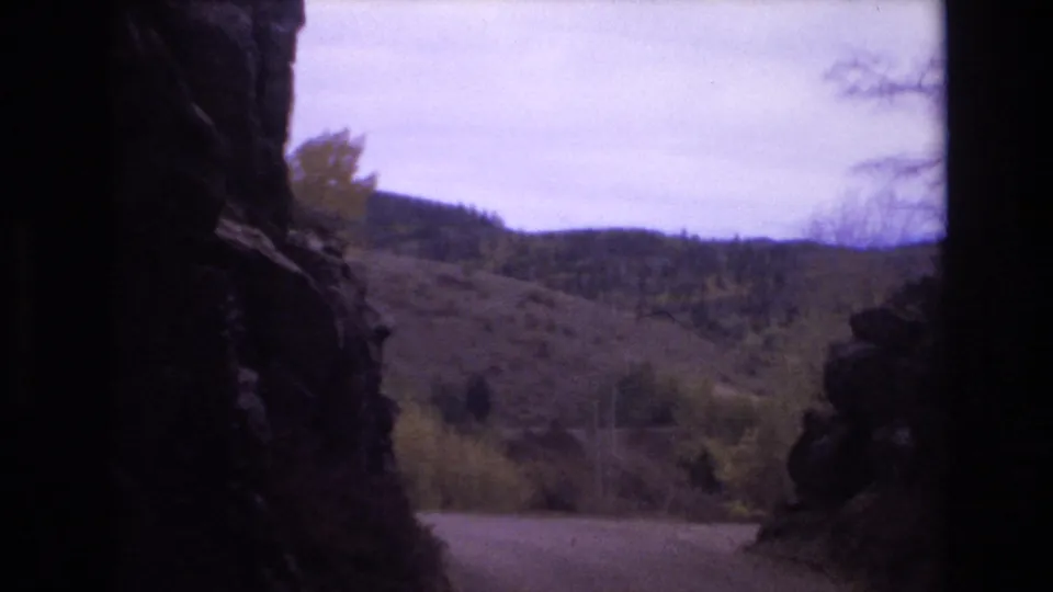 1973 Colorado Springs home-movie frame showing a winding roadside view between shadowed rock edges, with brush, trees, and low mountain ridges beyond.