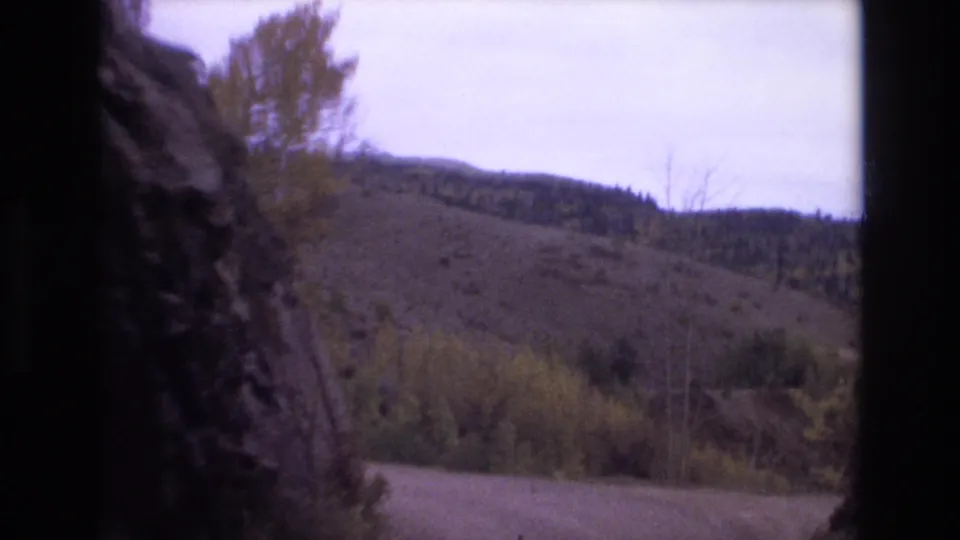 1973 Colorado Springs 8mm frame of a curving road below sparse autumnal trees and rolling hills, with a dark rock face along the left side.