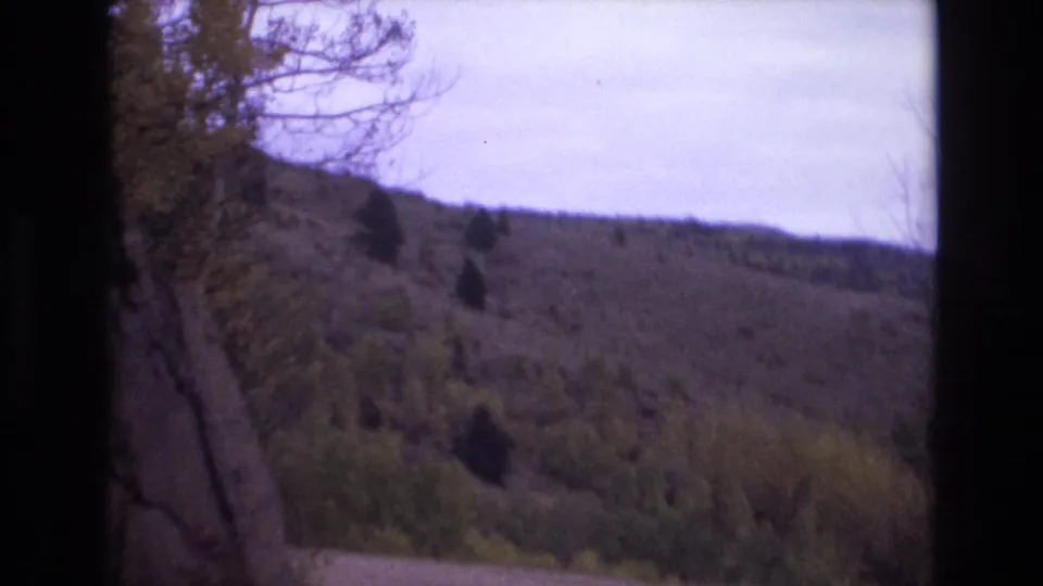 1973 Colorado Springs home-movie frame showing a dark vertical rock edge beside a brushy hillside, scattered trees, and a pale overcast sky.