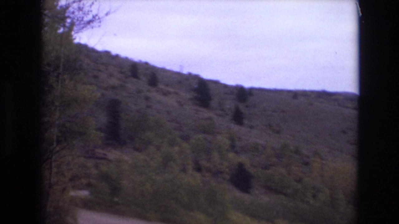 1973 Colorado Springs home-movie frame looking toward a winding dirt trail below a sparse grassy hillside with scattered evergreens and a pale sky.