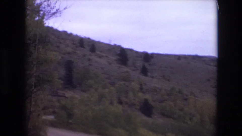 1973 Colorado Springs home-movie frame looking toward a winding dirt trail below a sparse grassy hillside with scattered evergreens and a pale sky.