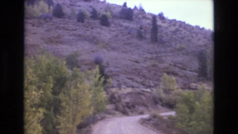 1973 Colorado Springs home-movie still with a dirt road bending past leafy shrubs beneath a sparse rocky hillside and scattered trees.