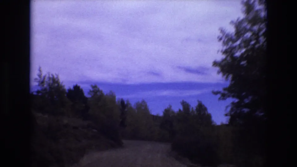 1973 Colorado Springs frame with a darker moving view along a dirt road lined by trees and shrubs beneath a blue, clouded sky.