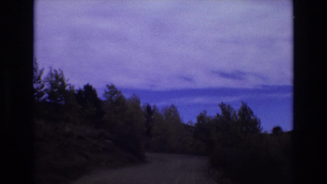 1973 Colorado Springs home-movie frame showing a winding dirt road through wooded hills under a cloudy blue-purple sky.
