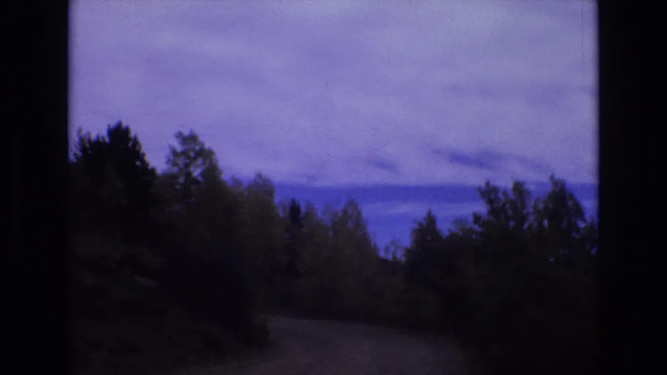 1973 Colorado Springs frame from a moving 8mm camera, with a curving road partly obscured by trees and hillside vegetation.