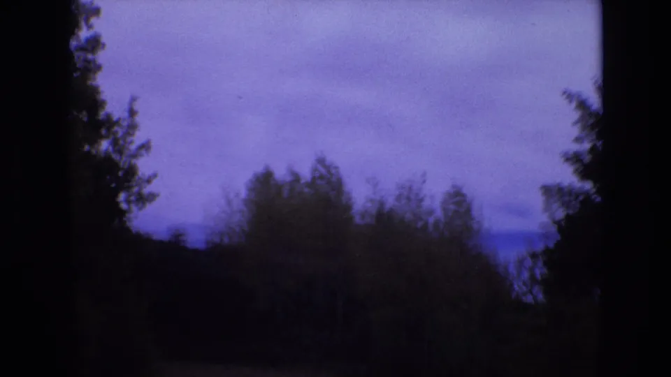 1973 Colorado Springs home-movie frame with dark roadside trees, wooded hillside, and cloudy sky as the road falls mostly out of view.