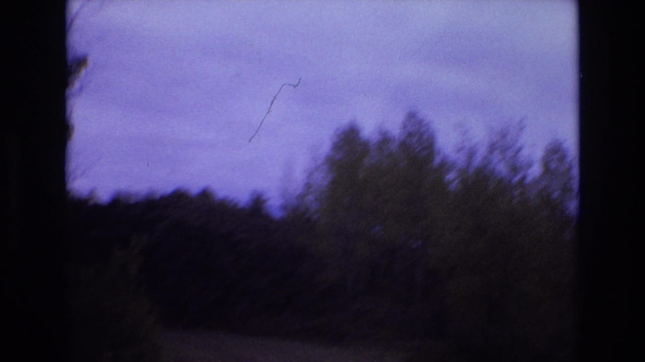 1973 8mm home-movie frame from the Colorado Springs clip showing a dark mountain roadside with brush, distant tree line, and a purple-blue sky framed by black gate edges.