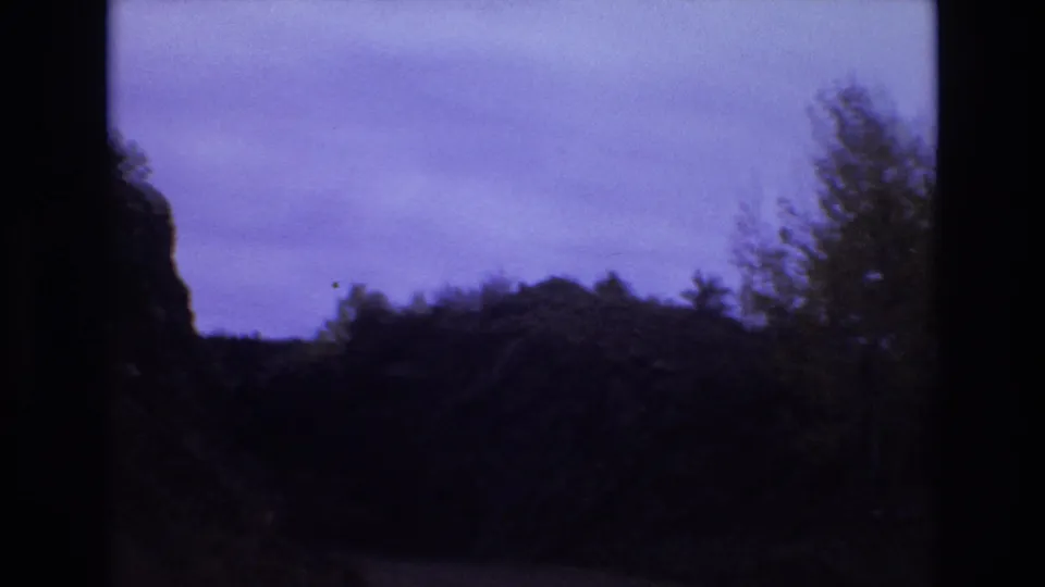 1973 Colorado Springs 8mm frame showing the forward view into a rocky roadside cut with dark vegetation, uneven cliff edges, and a pale purple sky above.